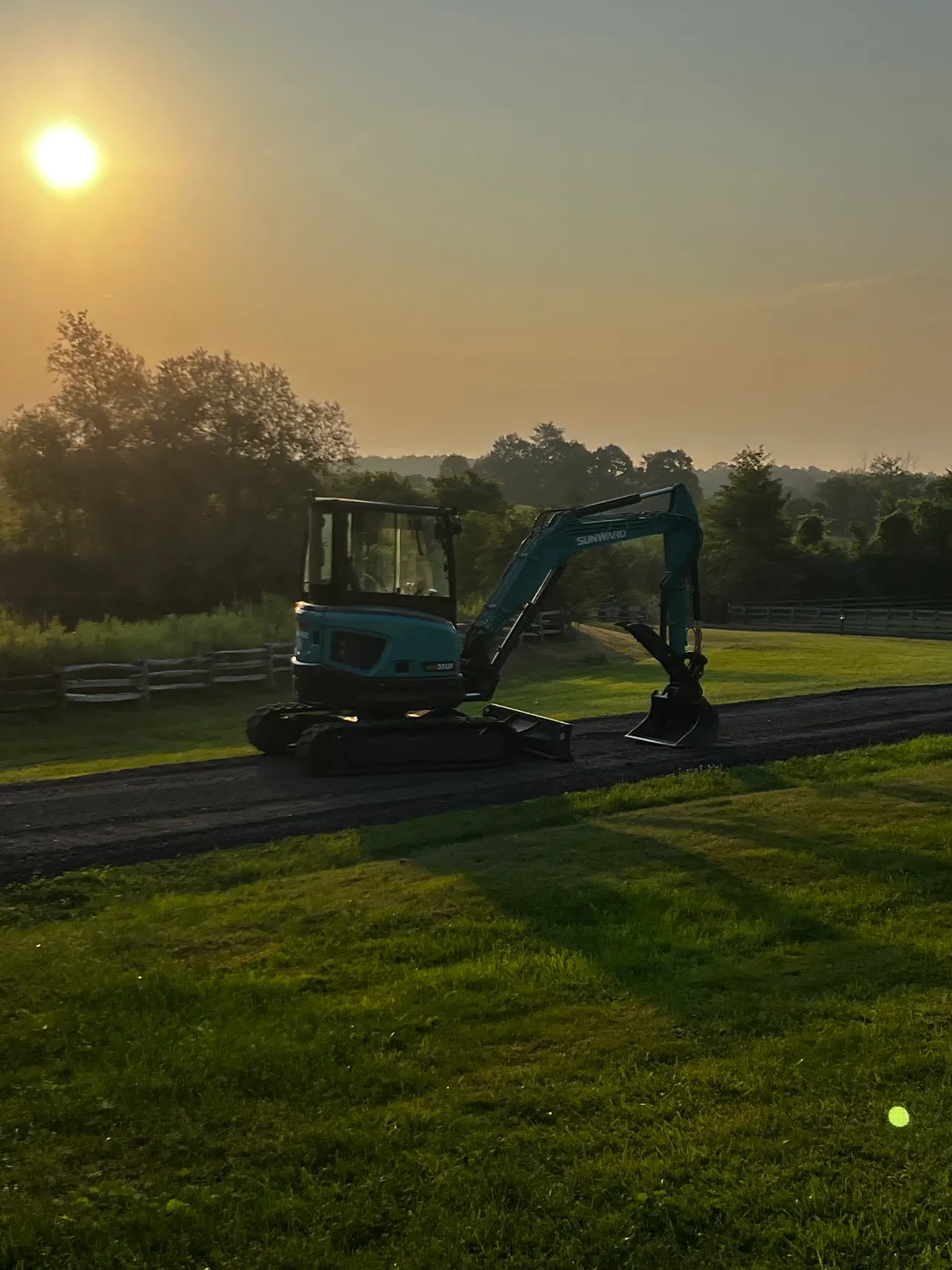 A mini excavator parked on a finished gravel laneway beside a white estate fence at golden hour