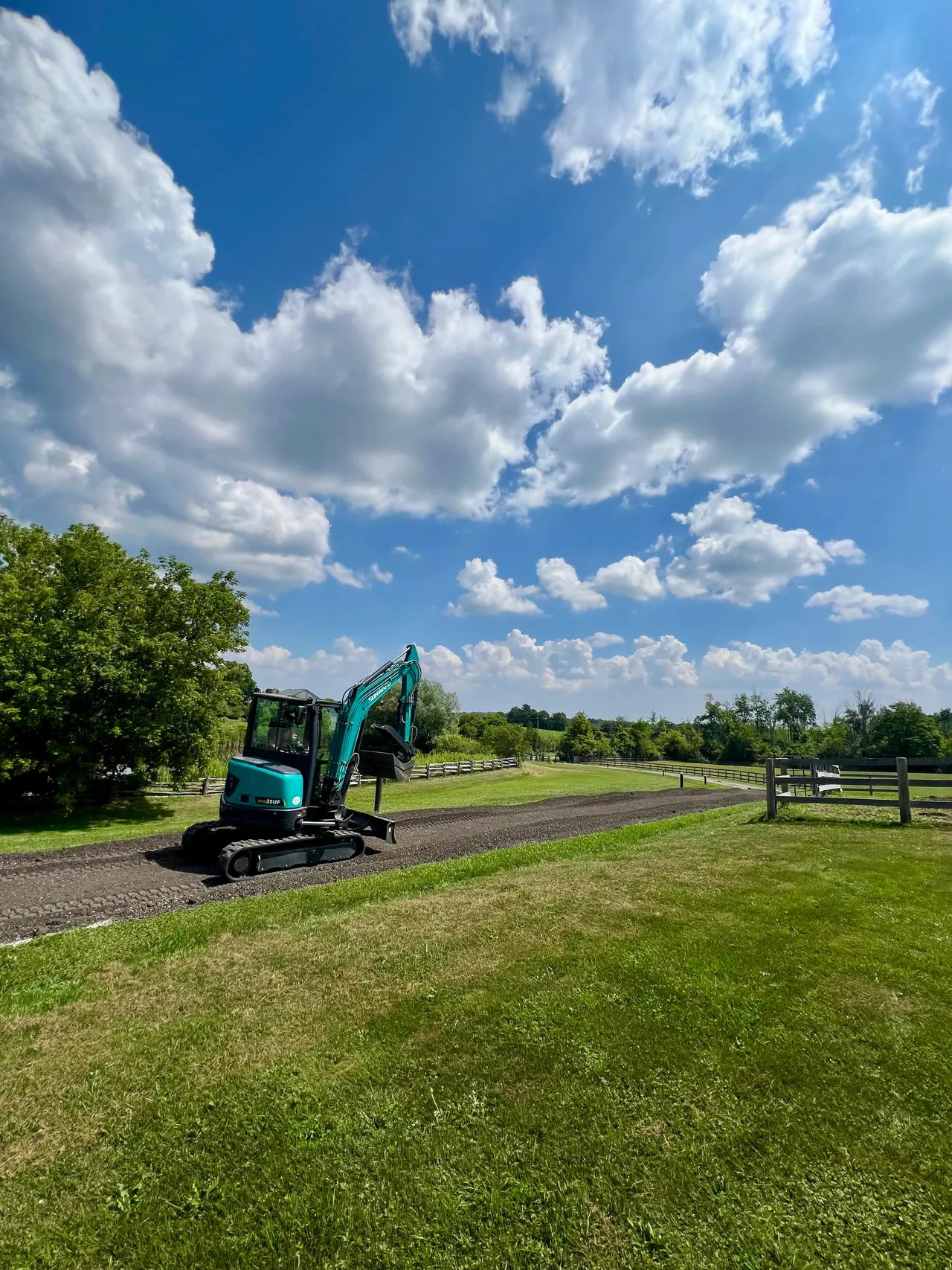 A mini excavator on a gravel laneway lined with white fences under a bright sky