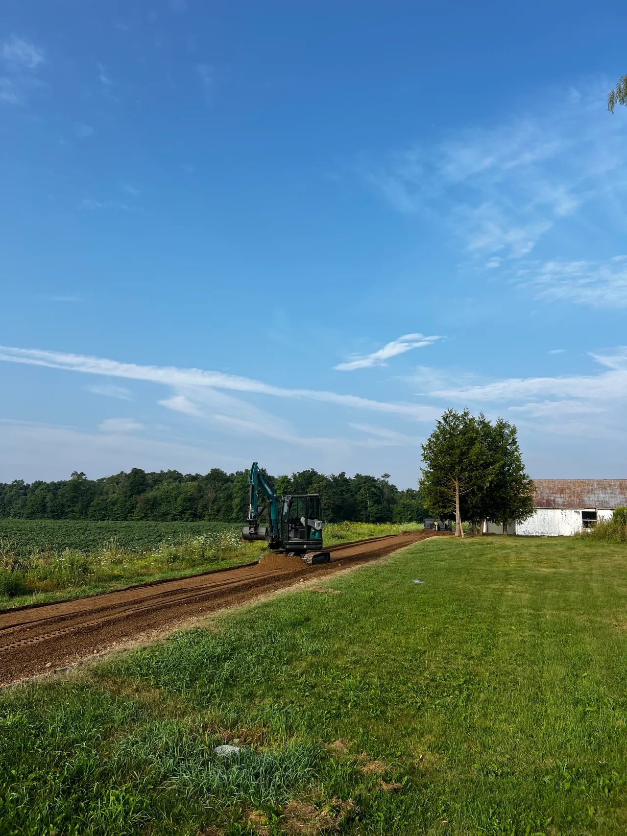 A mini excavator grading a new laneway beside a crop field and barn under a blue sky